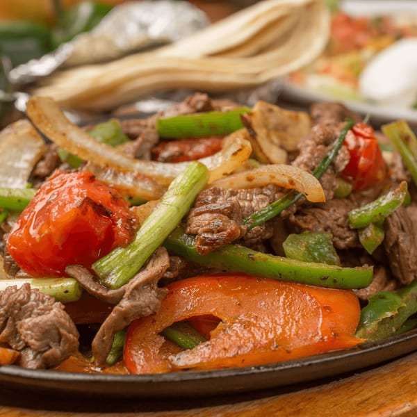 El Vaquero's sizzling fajitas with steak, bell peppers, onions, and tomatoes, with tortillas in the background.
