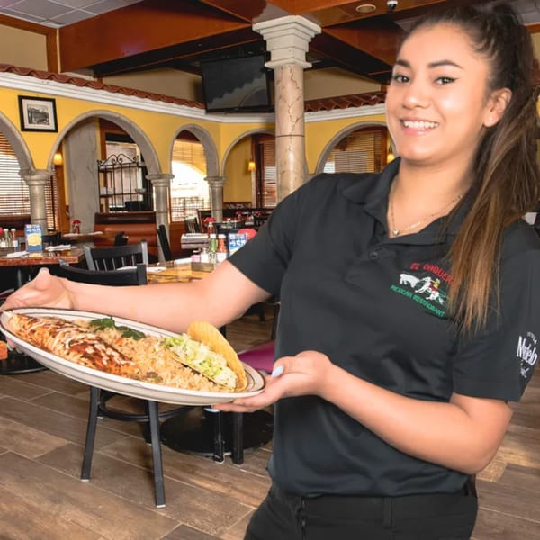 Smiling El Vaquero server holding a platter with a burrito, rice, and taco in a restaurant.