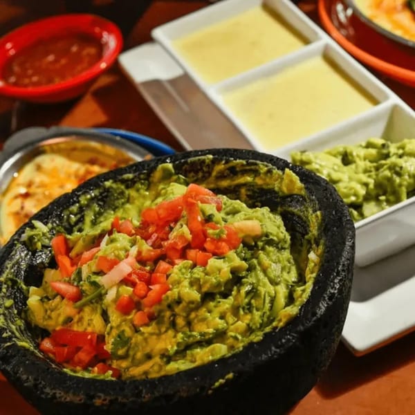 Fresh guacamole with diced tomatoes and onions in a black molcajete, alongside queso dip and salsa.