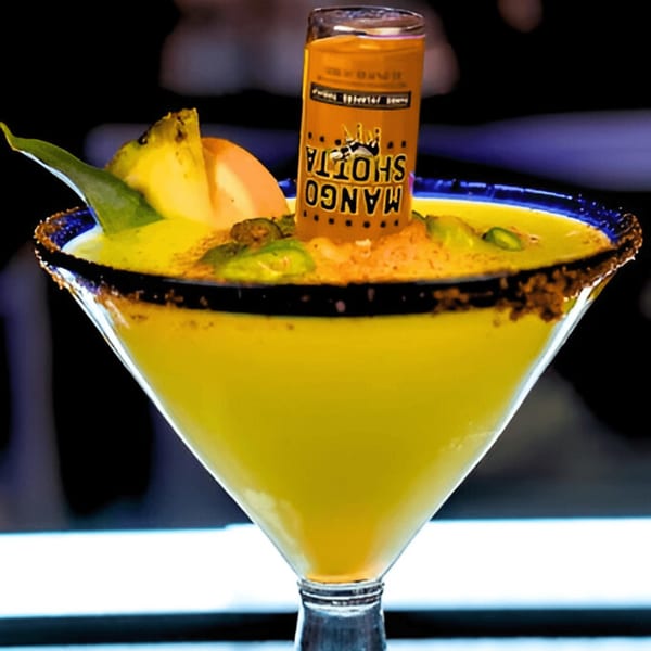 A mango margarita with a "Mango Shotta" shot glass inverted in it, garnished with fruit and chili.