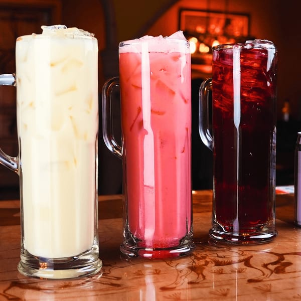 Three tall glasses of horchata, pink agua fresca, and red agua fresca on a wooden table.