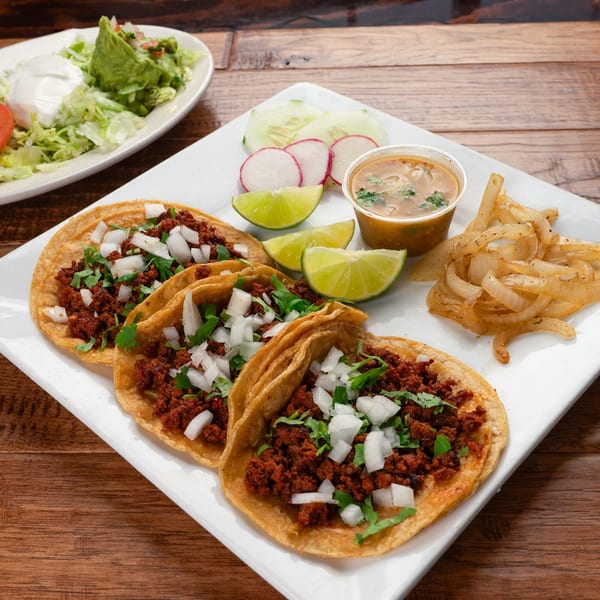Three El Vaquero chorizo tacos with onions and cilantro, served with limes, radishes, and a side of soup.