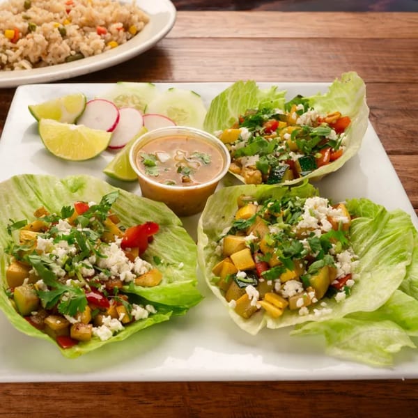 Three lettuce wraps filled with vegetables and cheese, served with lime, radish, cucumber, and sauce.