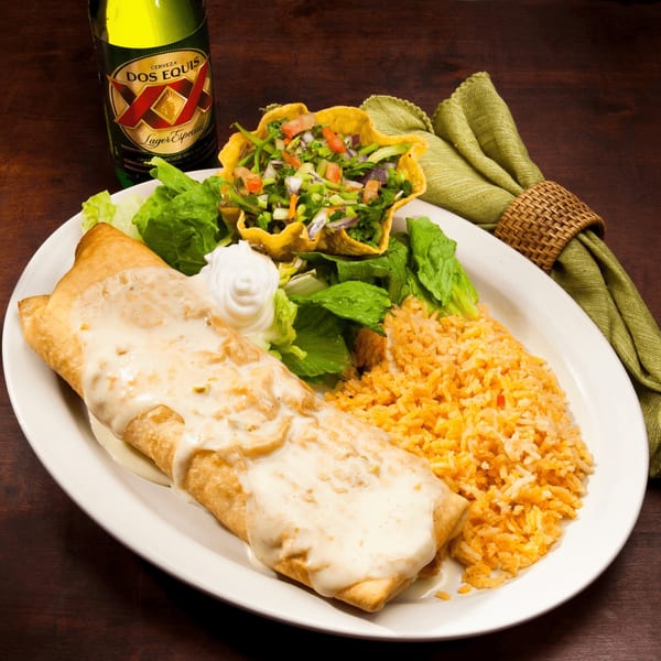 A chimichanga with white sauce, rice, salad in a taco shell, and a Dos Equis beer.