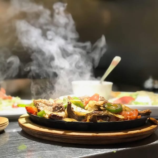 Sizzling beef fajitas with peppers and onions on a cast iron platter, emitting steam.