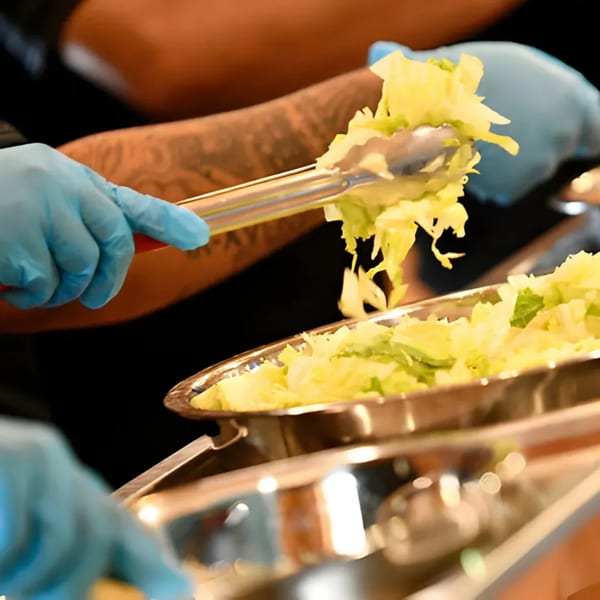 Gloved hands using tongs to serve shredded lettuce from a metal tray at El Vaquero.