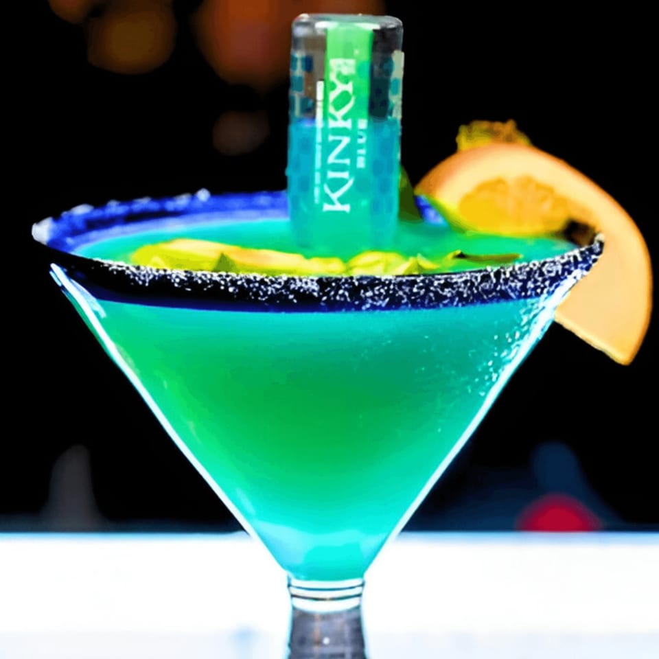 A vibrant blue-green margarita in a martini glass, garnished with a Kinky Blue shot and orange slice.