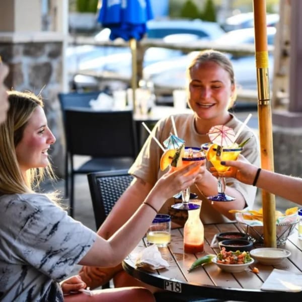 Three women clinking festive margaritas at an outdoor El Vaquero restaurant table with chips and salsa.